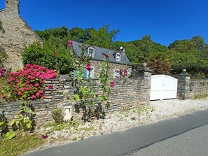 A stone house with a white gate, surrounded by lush greenery and flowering plants. - Charming house with fireplace and private garden (Surtainville)