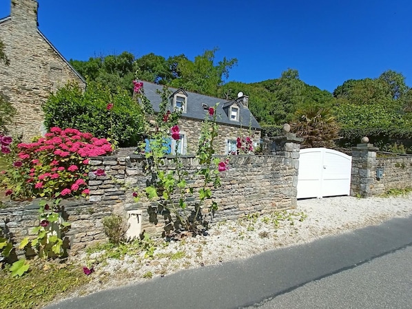 A stone house with a white gate, surrounded by lush greenery and flowering plants.