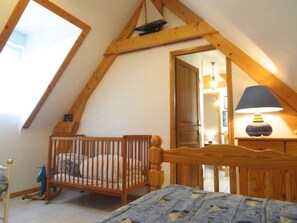 A bedroom with a sloped ceiling, a wooden crib, a bed with a blue patterned bedspread, and a lamp on a bedside table.
