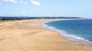 A wide sandy beach with gentle waves, a clear sky, and distant hills.