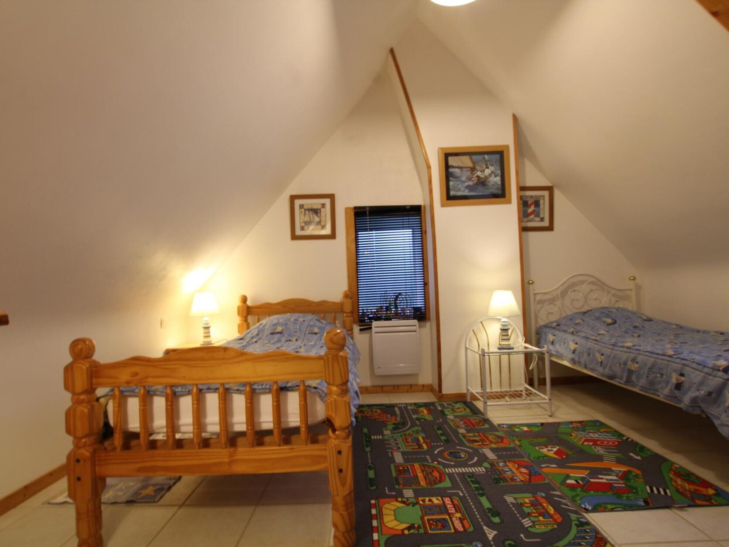 Een slaapkamer met twee houten bedden, een raam met zonwering en een kleurrijk speeltapis op de vloer.