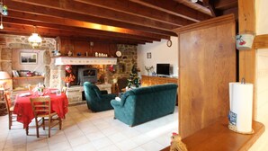 Un salon chaleureux avec une cheminée en pierre, des poutres apparentes, un coin repas avec une nappe rouge et un sapin de Noël.