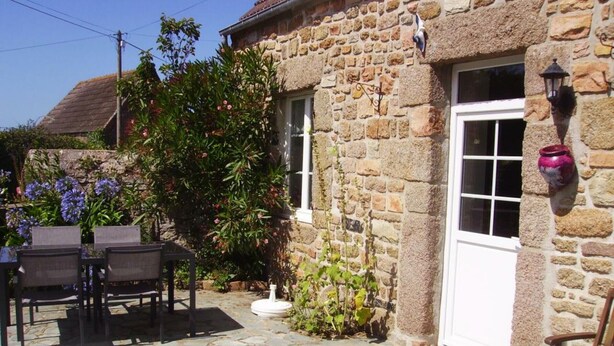 Une maison en pierre avec une porte blanche, un petit jardin fleuri et une terrasse en pierre.