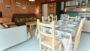 Un salon chaleureux avec un plafond en bois, un canapé, une table à manger pour quatre personnes et un coin cuisine équipé de placards et d’un évier.
