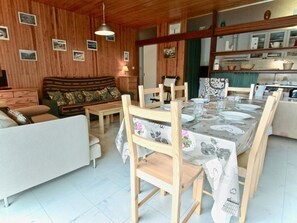 A cozy living room with a wooden ceiling, a sofa, a dining table set for four, and a kitchen area with cabinets and a sink.
