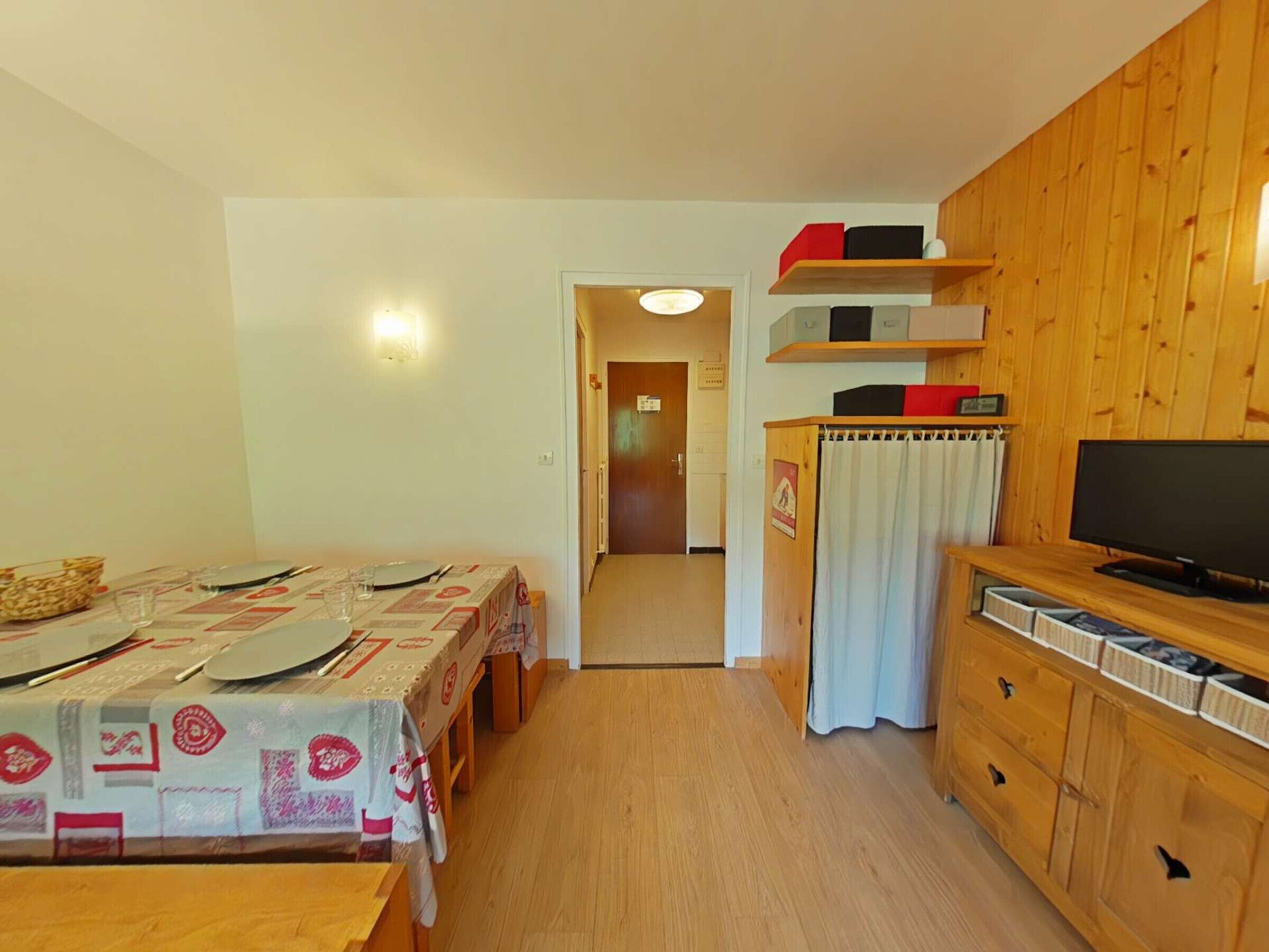 A small, cozy room with a wooden table set for two, a television, and a cabinet with storage drawers.