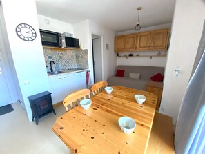 A compact kitchen with a microwave, a stove, and a sink. There is a dining table with four chairs and two bowls. The room has wooden cabinets and a clock on the wall.