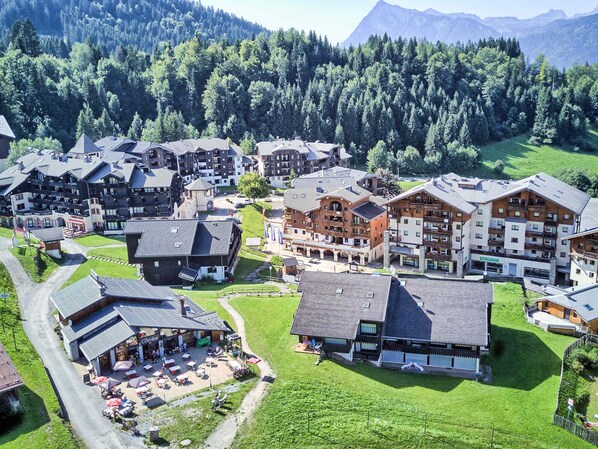 A mountainous village with traditional chalet-style buildings, a central square with outdoor seating, and a mountain backdrop.