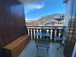 A balcony with a wooden bench and a table with chairs, overlooking a residential area with houses and a mountain.