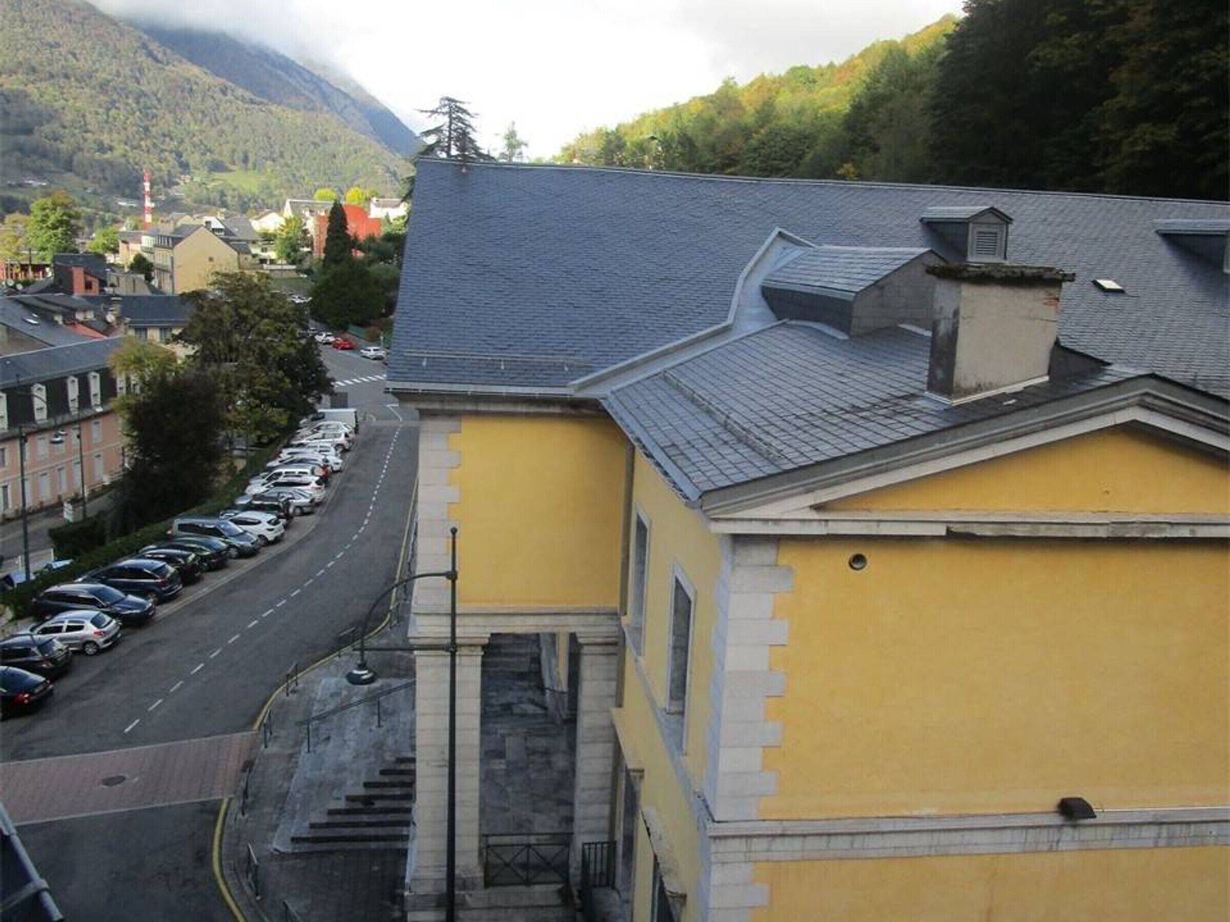 A yellow building with a grey roof, a parking lot with cars, and a mountainous background.