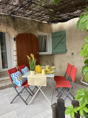 Eine Außenpatio mit einem Tisch für zwei Personen, umgeben von roten Stühlen, einer gelben Vase mit Blumen und einer grünen Tür.