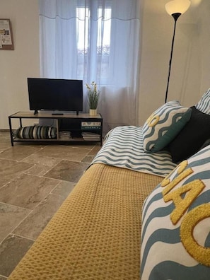 Olohuone, jossa on sohva, jalustalla oleva televisio, lattiavalaisin ja ikkuna, jossa on verhot.