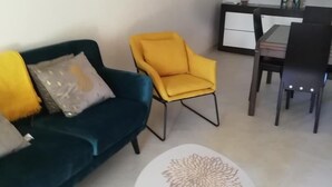 Een woonkamer met een donkerblauwe bank, een gele fauteuil en een kleine witte tafel met een decoratief middelpunt. Achtergrond toont een keuken met een eettafel en stoelen.