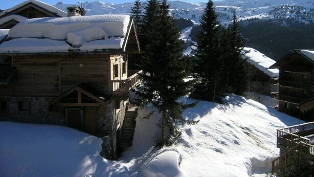 Una cabaña de madera con techo nevado, una escalera que lleva a la entrada y una vista de montañas cubiertas de nieve al fondo.