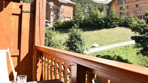 Un balcón de madera con una mesa y sillas, con vistas a un área verde con edificios y árboles al fondo.