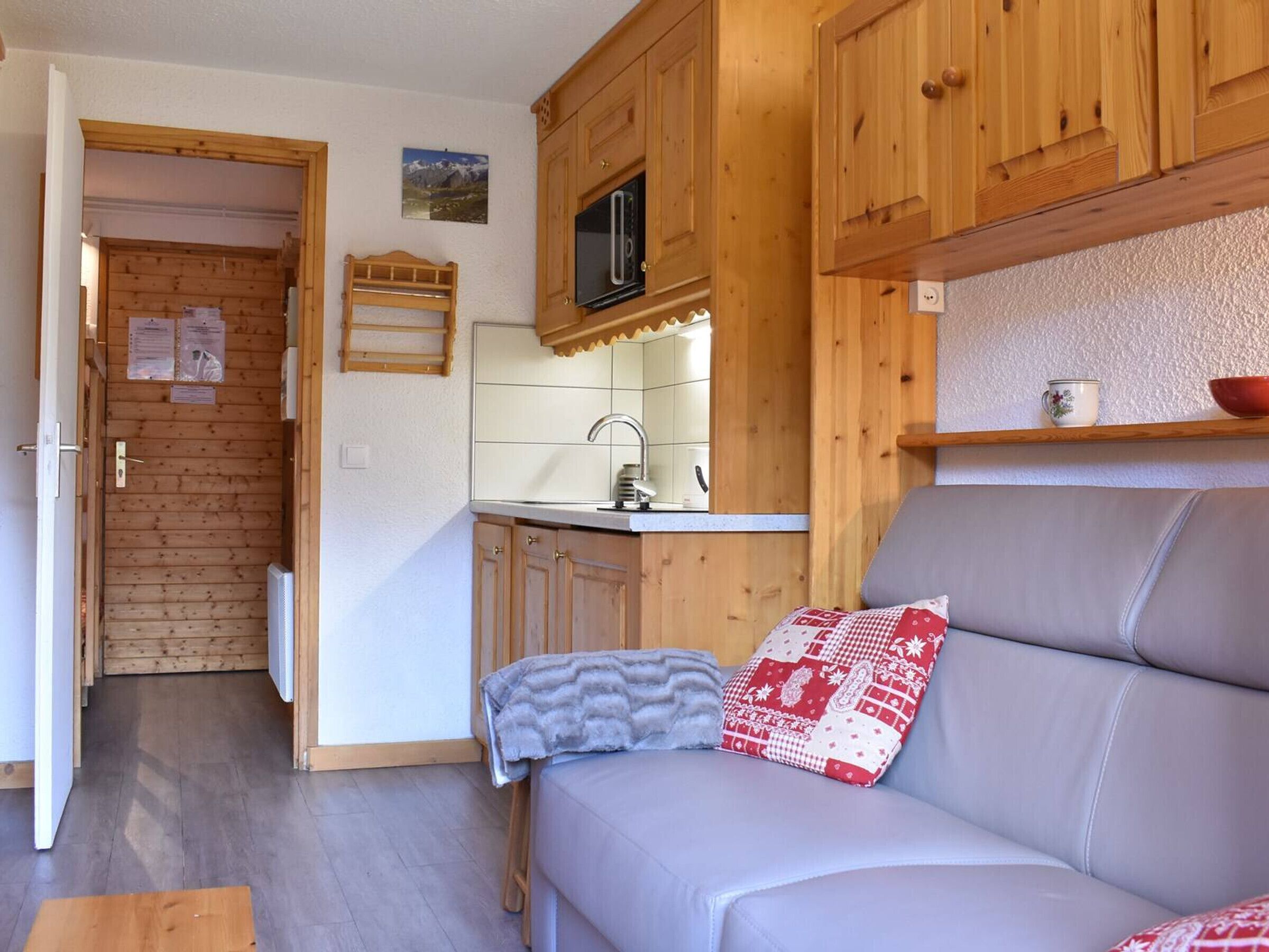 A compact living space with a kitchenette, a sofa with a patterned cushion, and a wooden door leading to a bathroom.