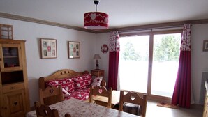 A cozy room with a wooden bed, a dining table with chairs, a lamp, and a window with curtains. - D002 - 60 m2 - 3P+cab - 6/8 pers (Prapoutel)
