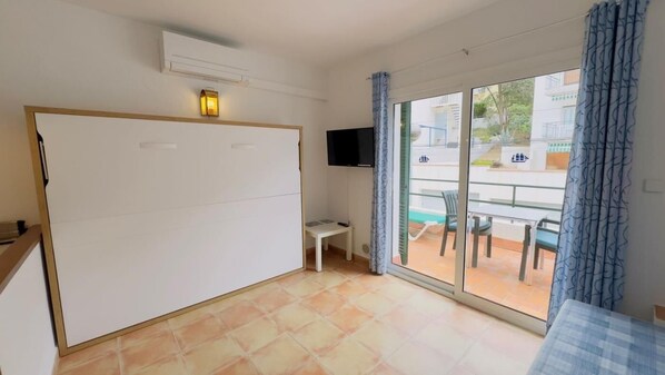 Ein Zimmer mit einer Schiebetür, die auf einen Balkon führt, einem Wand-Flachbildfernseher und einem kleinen Tisch mit Stuhl.