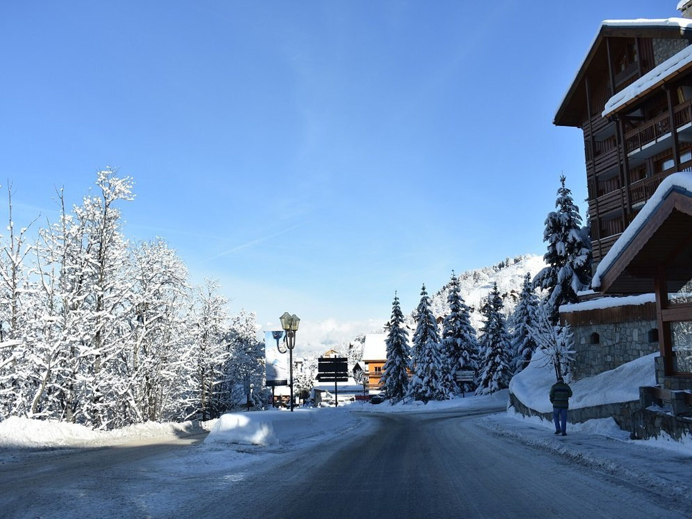 A snow-covered road leading to a wooden building with a balcony, surrounded by snow-laden trees and a clear sky.
