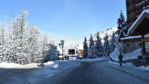 A snow-covered road leading to a wooden building with a balcony, surrounded by snow-laden trees and a clear sky.