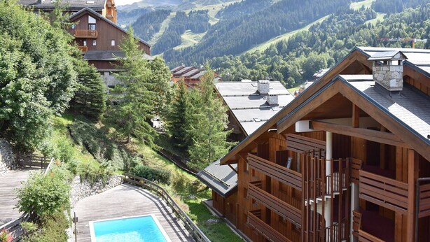 A wooden chalet with a wooden deck and a pool, surrounded by forested hills and mountains.