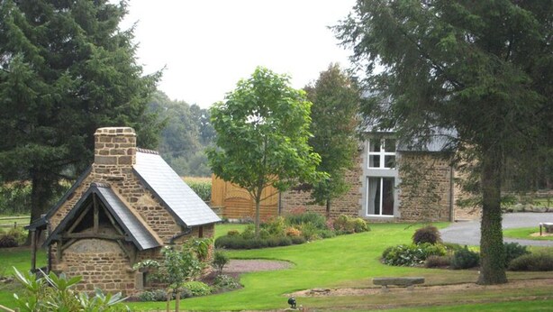 A stone cottage with a sloped roof, surrounded by a well-maintained lawn and trees. - Charming house 3 bdrms., garden, 6 pers. (Le Grand-Celland)