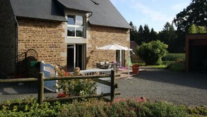 A stone house with a sloped roof, a front door, and a small porch with outdoor furniture. - Charming house 3 bdrms., garden, 6 pers. (Le Grand-Celland)