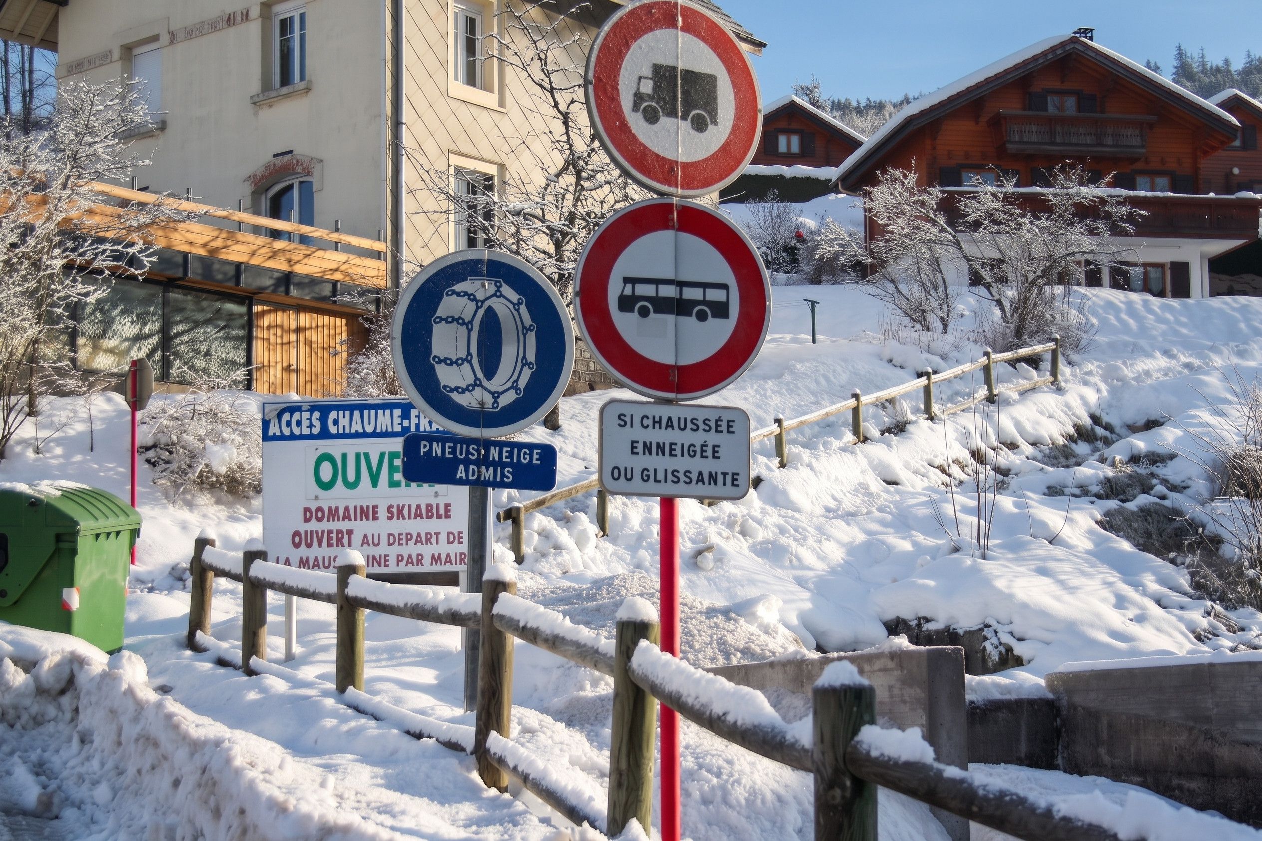 Une scène hivernale en plein air, avec une clôture en bois, une poubelle verte et plusieurs panneaux de signalisation.