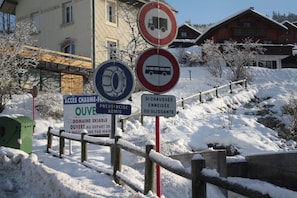 Ein verschneiter Außenbereich mit Holzzaun, einem Schild mit verschiedenen Symbolen und einem Gebäude im Hintergrund.