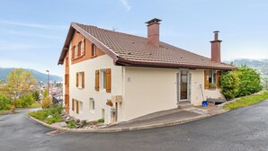 Ein zweistöckiges Haus mit geneigtem Dach und Schornsteinen, umgeben von einem Garten und einer Straße.