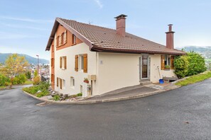 Ein zweistöckiges Haus mit geneigtem Dach und Schornsteinen, umgeben von einem Garten und einer Straße.