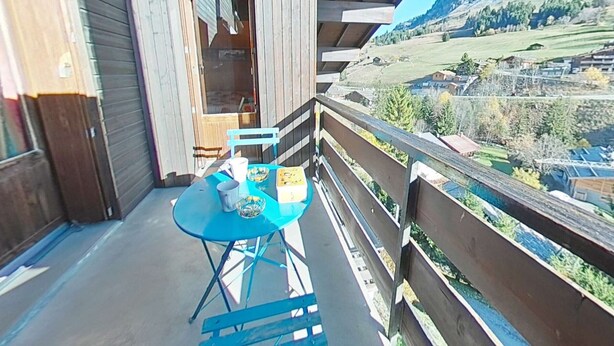 Un balcon en bois, agrémenté d’une table et de chaises, qui offre une vue sur une vallée parsemée de maisons et bordée de montagnes.