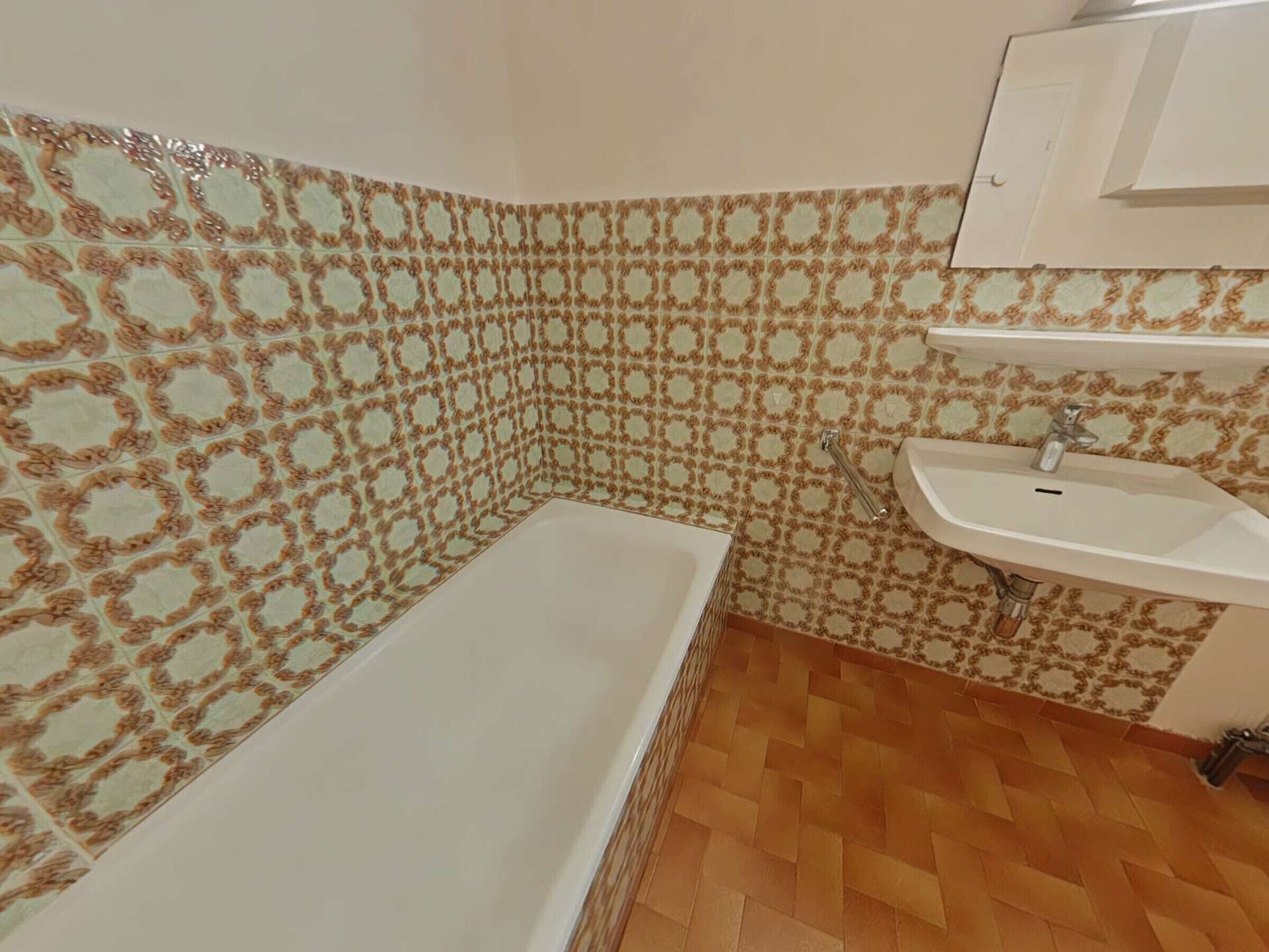 Une salle de bain avec un mur recouvert de carreaux à motifs, une baignoire blanche, un plancher en bois et un lavabo blanc avec un miroir au-dessus.
