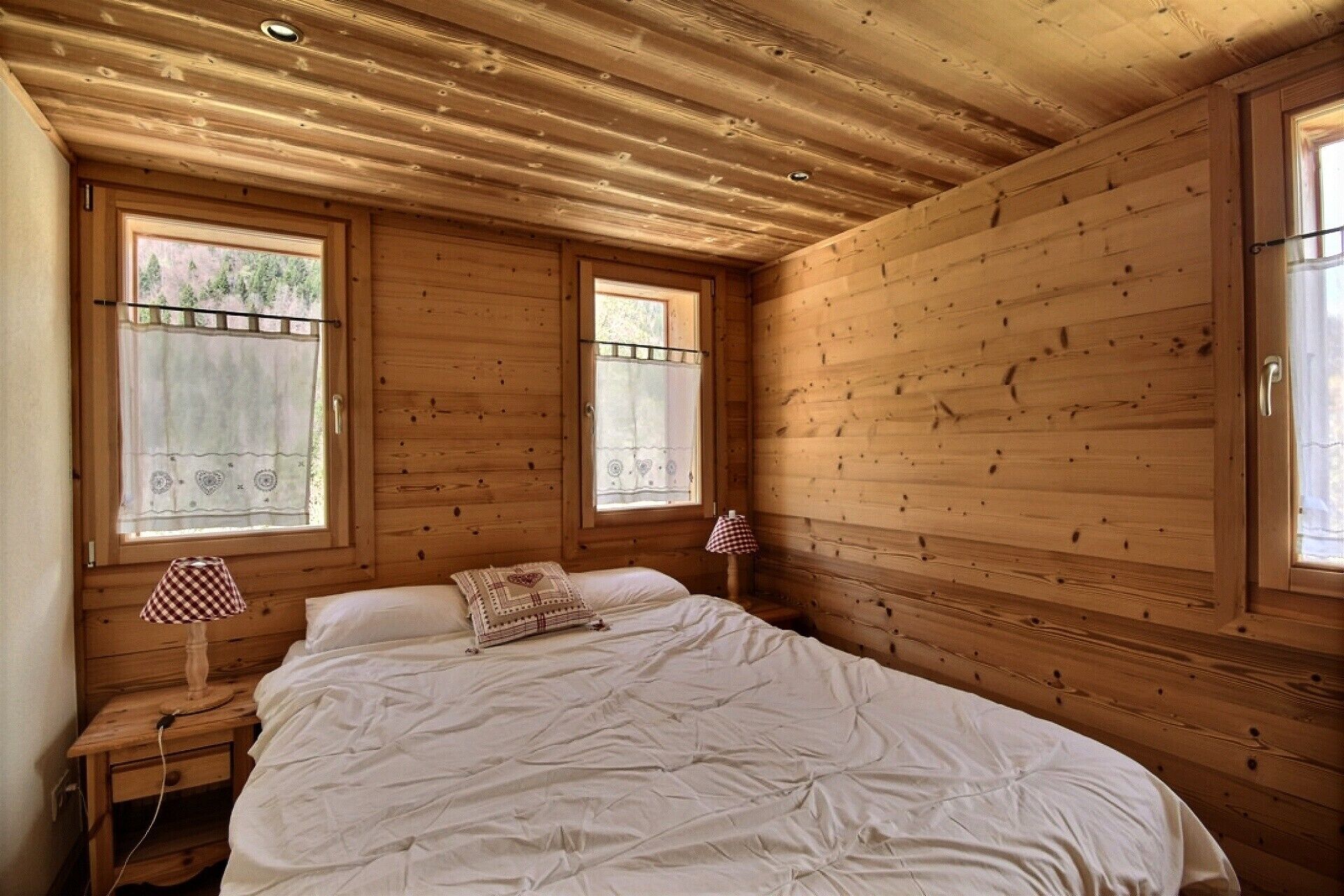 Ein Schlafzimmer einer Holzhütte mit einem Bett, zwei Fenstern und einem Nachttisch mit Lampe.