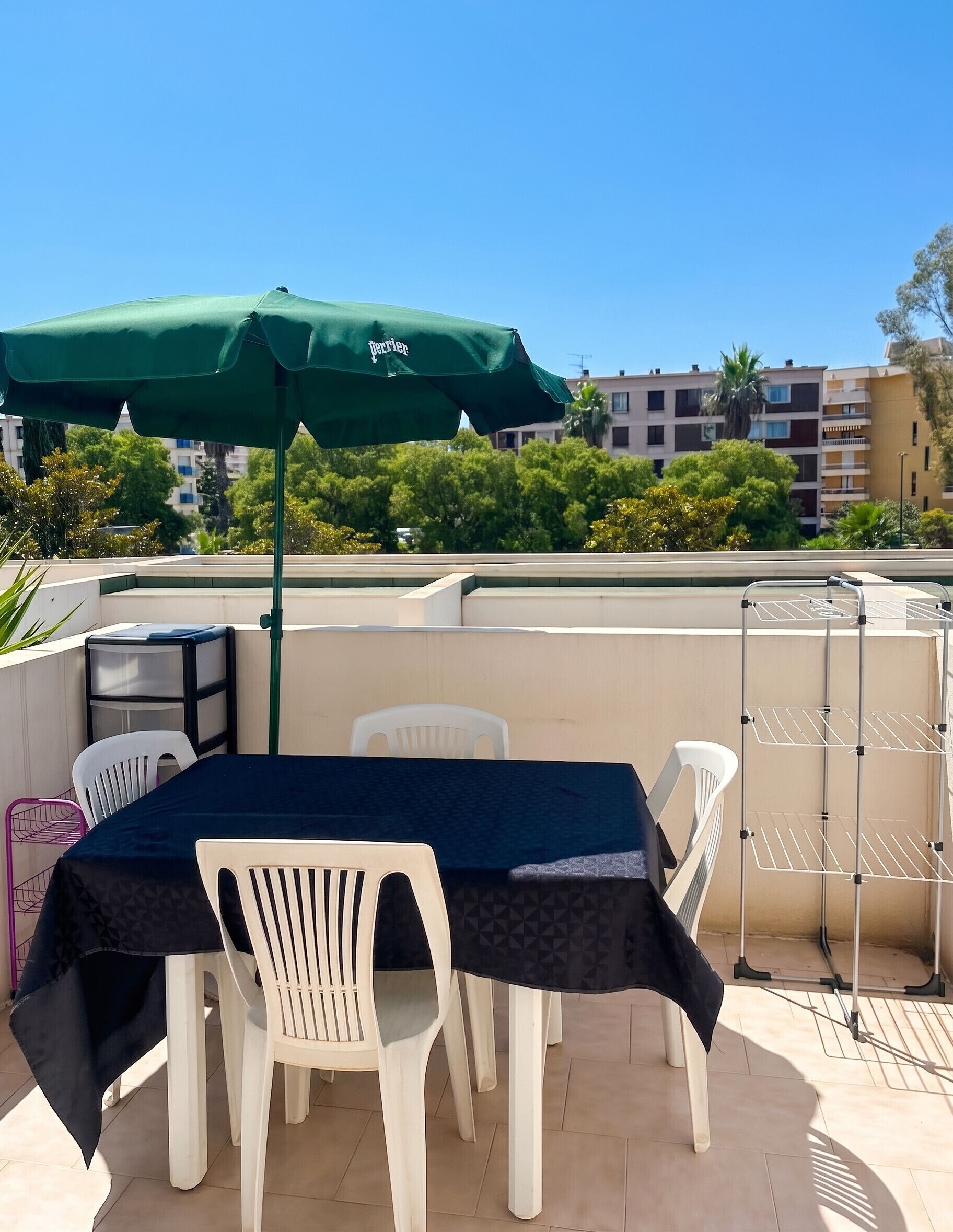 Een terras buiten met een tafel, stoelen, een groene parasol en een metalen wasrek.