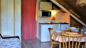 A cozy attic bedroom with a bed, a wooden dining table with chairs, a kitchen area with a microwave and a sink, and a red curtain.
