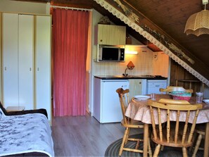 Um quarto de sótão acolhedor com cama, uma mesa de jantar de madeira com cadeiras, um espaço de cozinha com micro-ondas e pia, e uma cortina vermelha.
