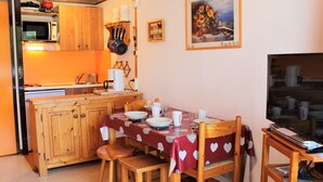 A compact kitchen with wooden cabinets, a microwave, and a sink. There is a dining table with chairs and a TV on a wooden stand.