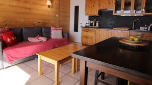 A cozy room with a wooden ceiling, a sofa with red and white cushions, a wooden table, and a kitchen area with wooden cabinets and a fruit bowl.
