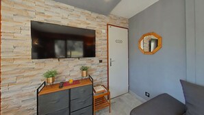 A room with a stone-patterned wall, a flat-screen TV mounted above a wooden desk with drawers, a small chair, and a bed with a gray blanket.