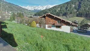 A chalet with a balcony, surrounded by lush greenery and a mountainous backdrop.