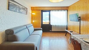 En hyggelig stue med en sofa, et tv ophængt på væggen og en trædør, der fører ud til en balkon.