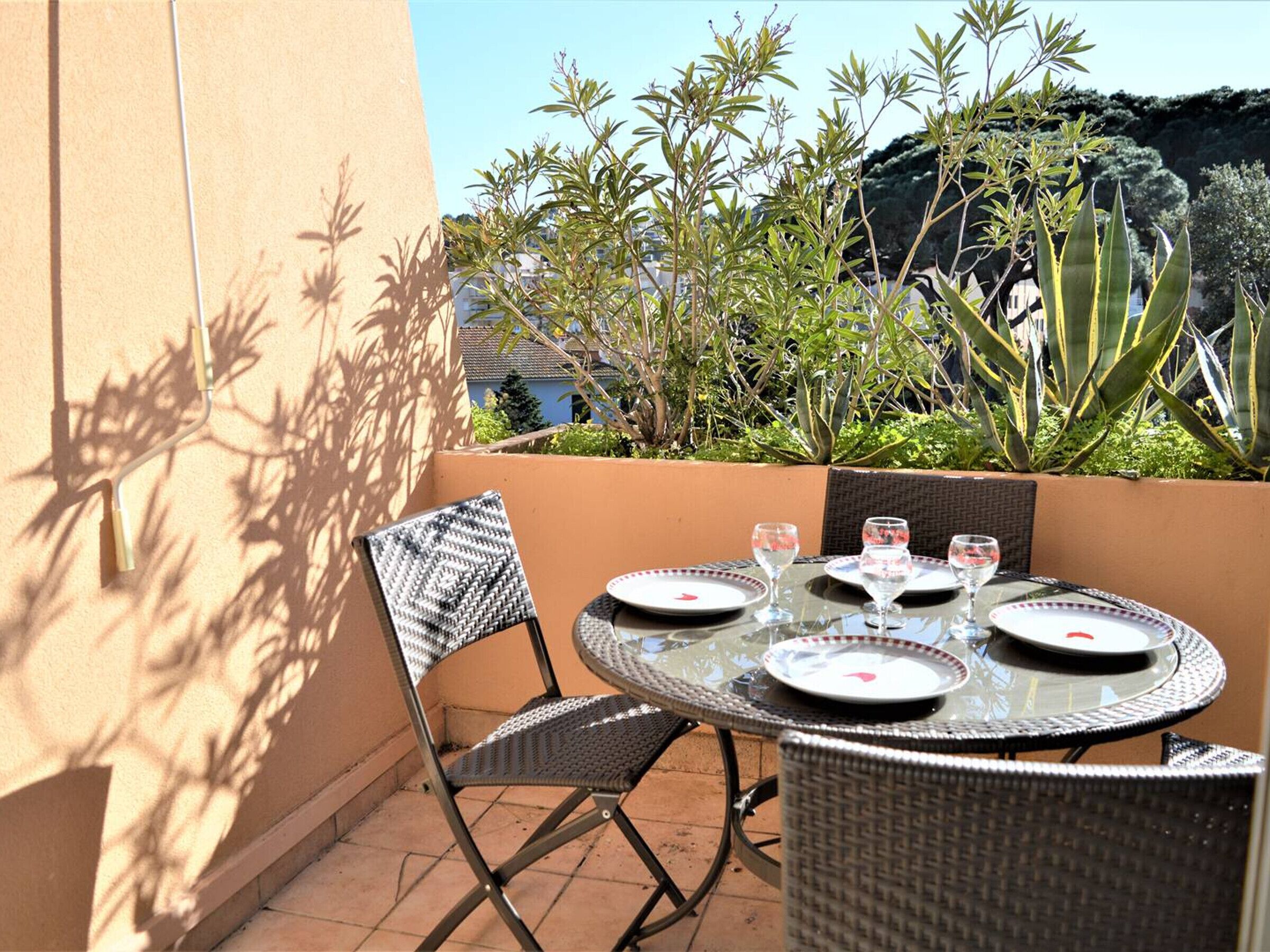 A round glass table with four chairs, plates, and glasses on a patio with plants and a view of a building.