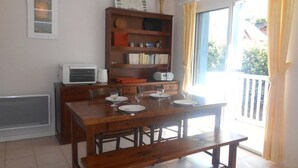 A dining area with a wooden table set for four, a bench, a bookshelf, and a window with curtains. - Cozy 3-Room Duplex with Balcony, Parking, Near Ski Lift, Sleeps 4 (Cauterets)