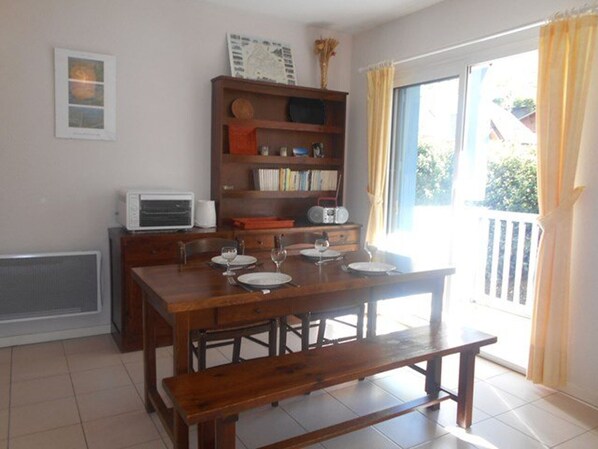 Uma sala de jantar com mesa de madeira posta para quatro pessoas, um banco, uma estante de livros e uma janela com cortinas.
