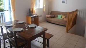A dining area with a wooden table set for two, a sofa with colorful cushions, a small desk with a lamp, and a window with curtains. - Cozy 3-Room Duplex with Balcony, Parking, Near Ski Lift, Sleeps 4 (Cauterets)