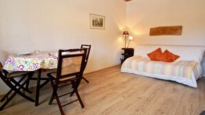 Une pièce avec un plafond en bois, une table à manger recouverte d’une nappe à motifs floraux, une chaise en bois, un lit avec une literie à rayures et une table de chevet avec une lampe.