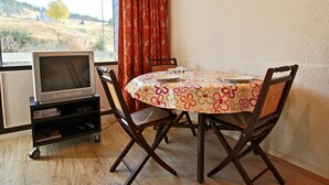 Un coin repas comprenant une table ronde, quatre chaises, un téléviseur posé sur un meuble et une fenêtre avec des rideaux.