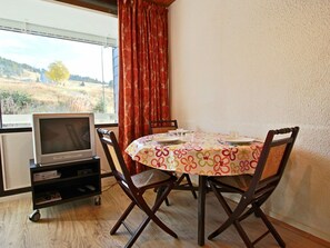 Un comedor con una mesa redonda, cuatro sillas, un televisor sobre un mueble bajo y una ventana con cortinas.
