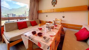 Ein Essbereich mit Tisch für zwei Personen, einer Couch mit roten Kissen und Blick auf Berge durch ein Fenster.